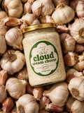 A glass jar of Cloud Cream Cheese Garlic, 400g, with a gold lid and green-and-white label, nestled among a dense pile of whole garlic bulbs.